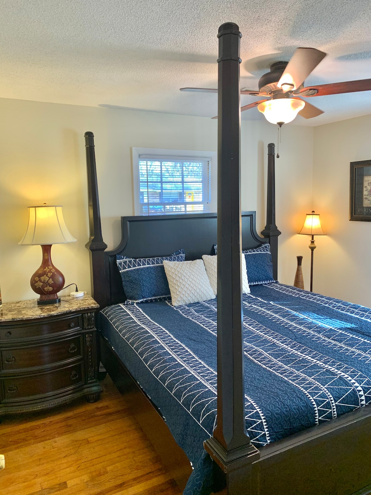 A four-poster bed with a navy blue quilt is positioned in a well-lit bedroom. A bedside table with a lamp is situated on one side, while a chest of drawers is on the opposite side. Natural light enters through a window adorned with blinds.