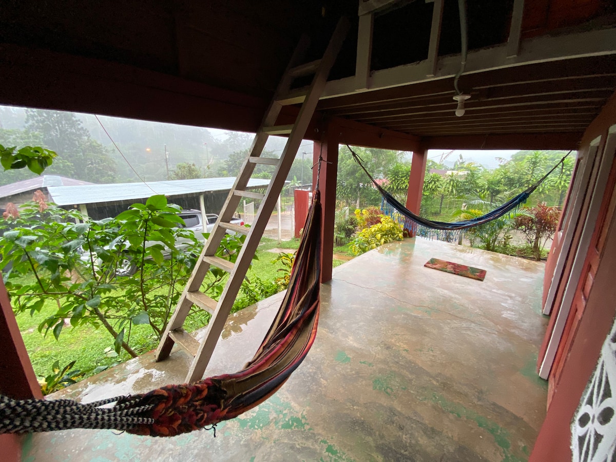 An outdoor patio area is revealed, featuring two colorful hammocks suspended between posts. A wooden ladder leans against the wall, leading to a lofted space. Lush greenery surrounds the area, with gentle rain creating a serene atmosphere.