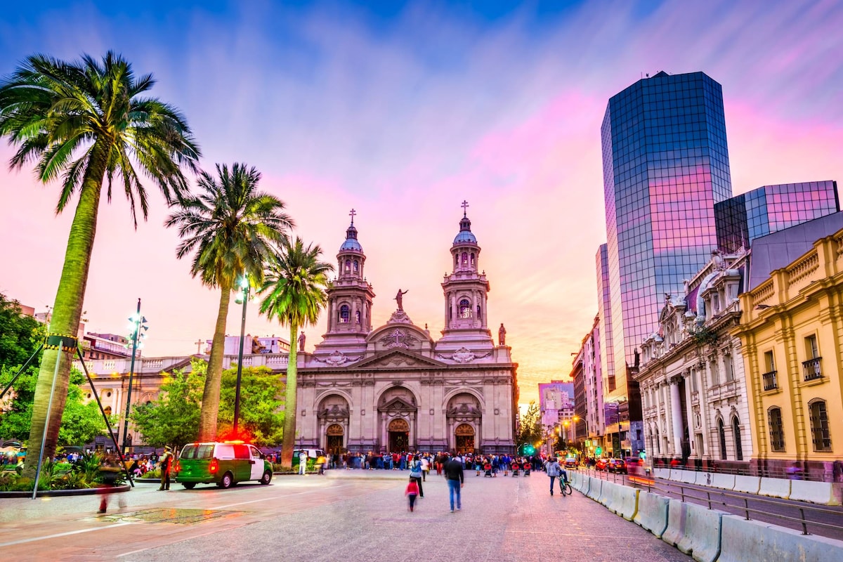 The image captures a bustling square in Santiago, featuring a historic church with twin spires, flanked by palm trees. Modern buildings rise in the background, and a diverse group of people can be seen enjoying the space during sunset, with vibrant colors reflecting in the sky.