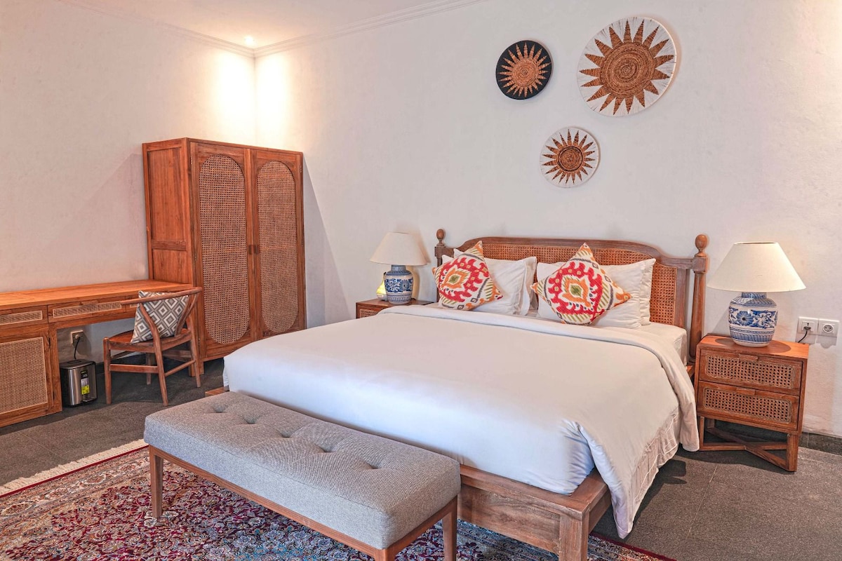A spacious room is adorned with a large bed, complemented by decorative cushions and a bench at the foot. Two bedside tables and a writing desk with a chair are present. Natural light fills the room, enhanced by decorative wall art and an elegant area rug.