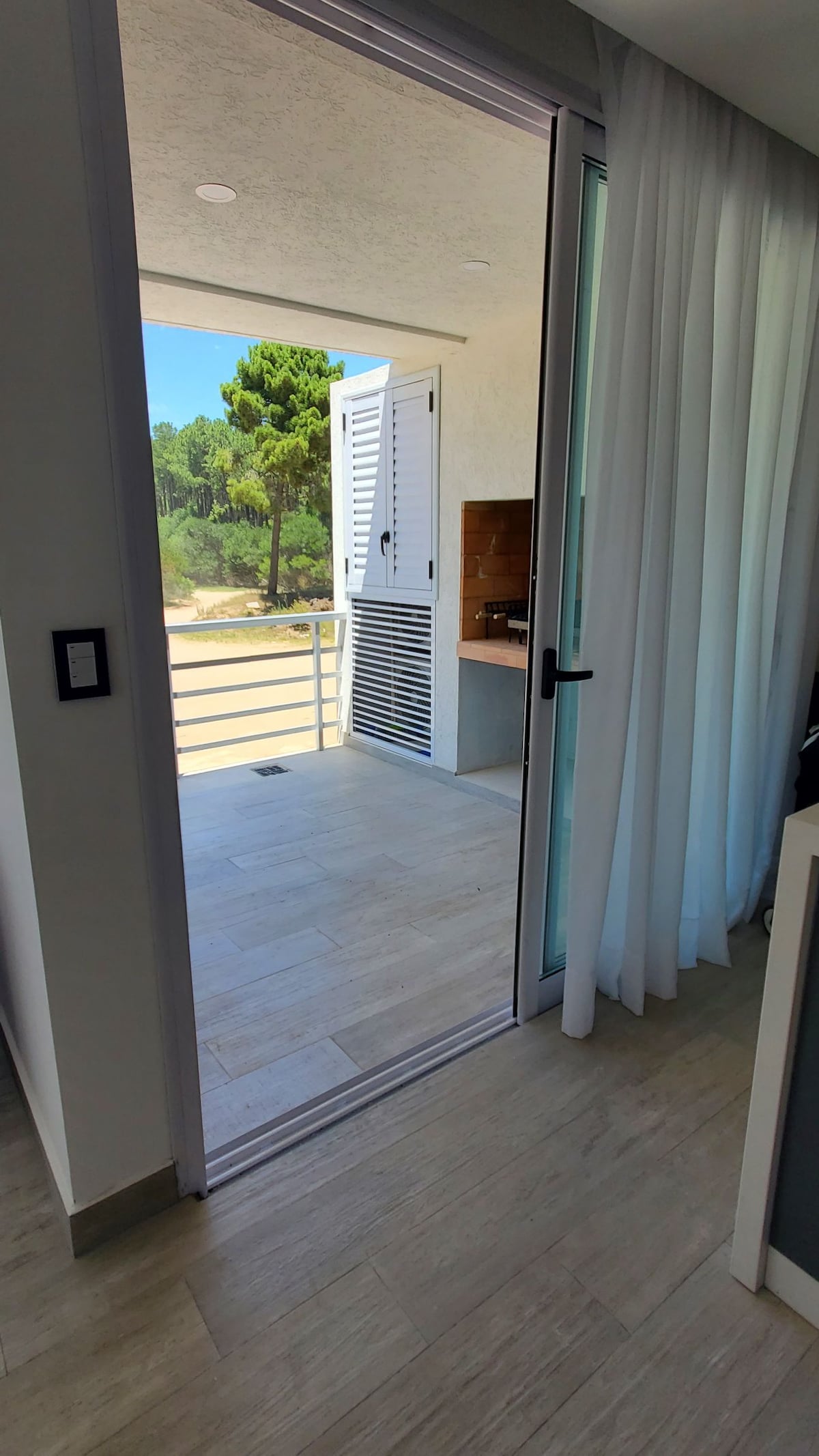 A view is offered from an open glass door leading to a balcony. Natural light enters the space, illuminating the neutral-toned flooring. The balcony features a grill and overlooks green trees in the distance, creating a connection with the outdoor environment.