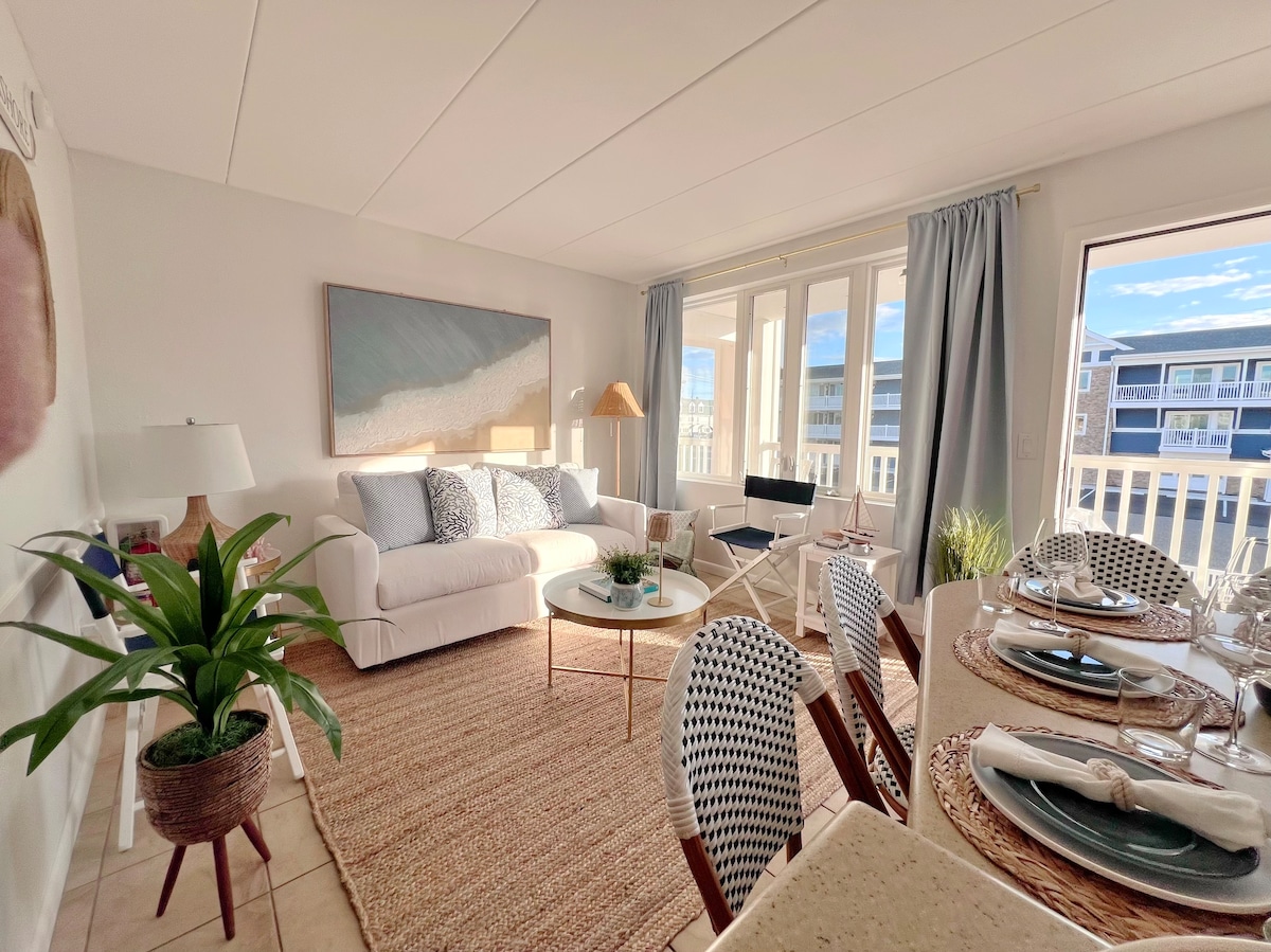 A bright living area features a white sofa adorned with blue and gray pillows, complemented by a glass coffee table. Natural light flows through large windows, revealing a view of the exterior. A dining table is set nearby, surrounded by patterned chairs.