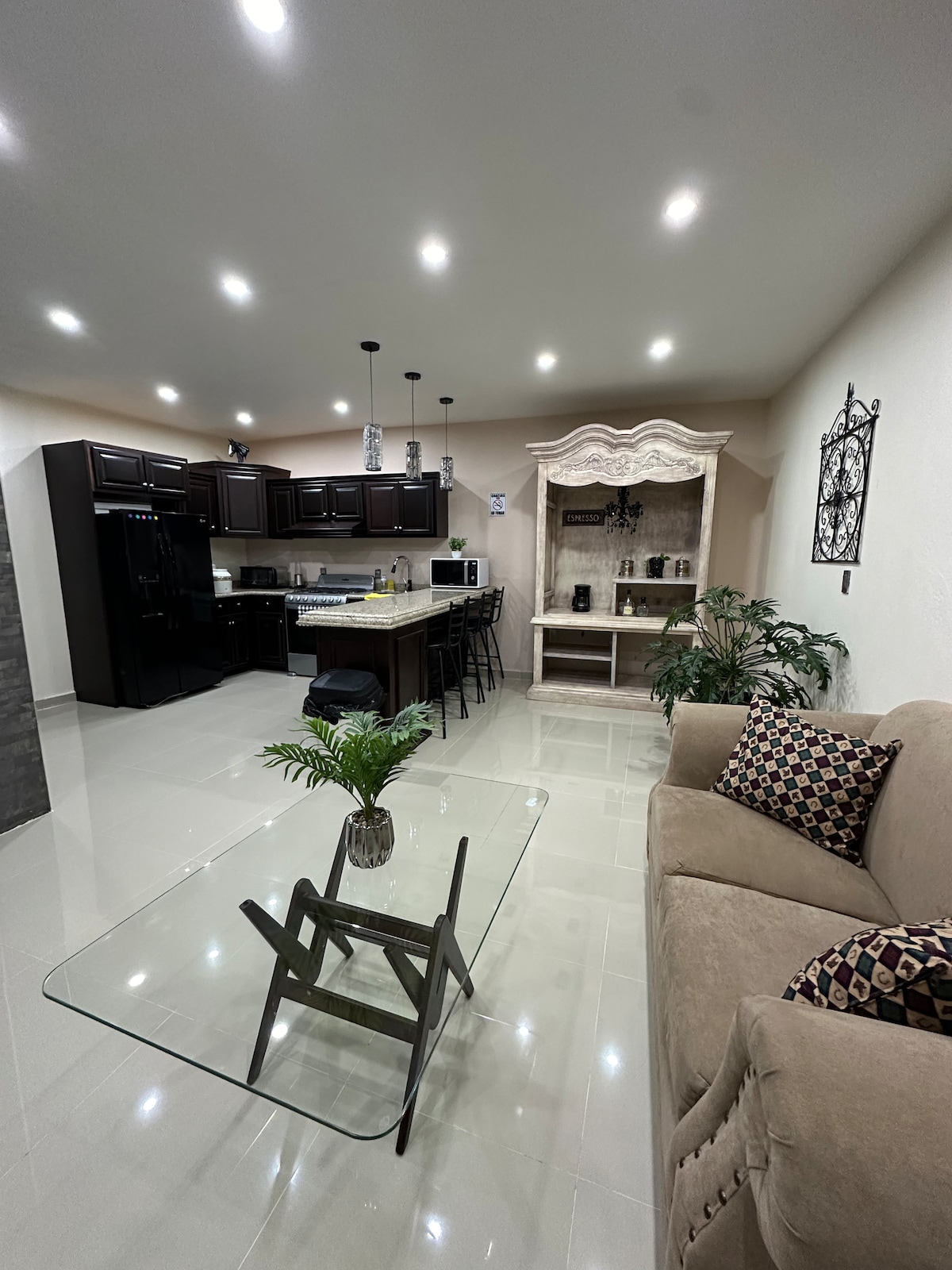 An open living area showcases a modern kitchen with dark cabinetry and stainless steel appliances. A glass coffee table is positioned atop polished tile flooring, next to a neutral-toned sofa adorned with patterned cushions. A decorative cabinet and potted greenery add to the elegant ambiance.