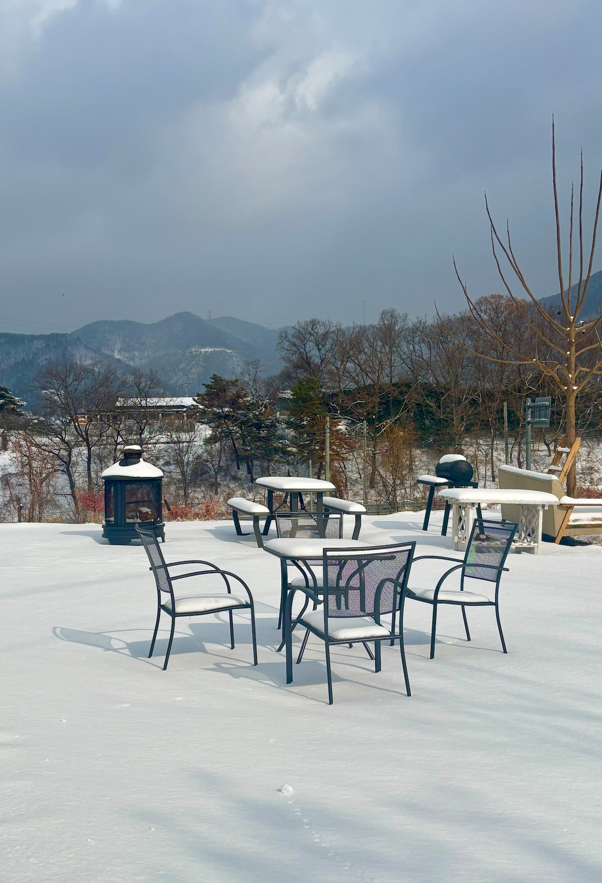 A wintery outdoor space features a round table surrounded by four black chairs atop a layer of fresh snow. In the background, dark mountains are partially obscured by clouds, while a grill and a lantern are visible, adding to the serene atmosphere.