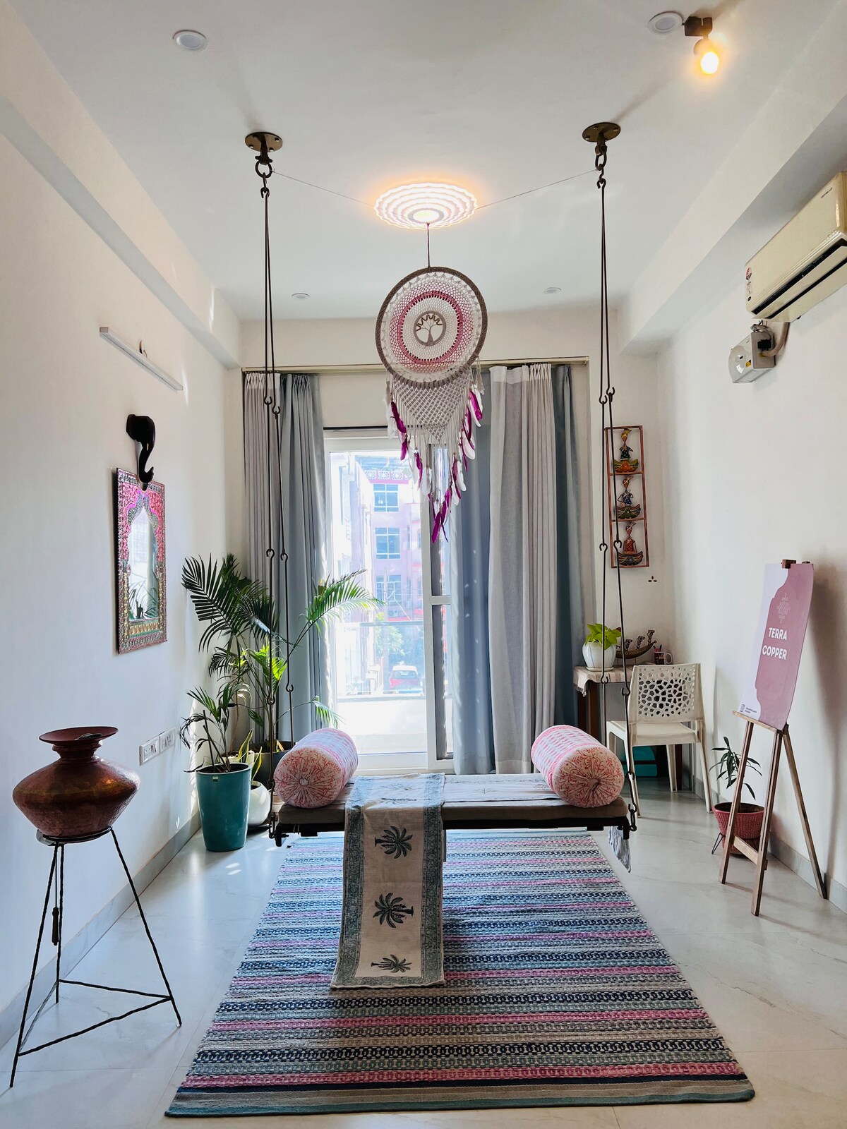 A spacious living area is illuminated by natural light coming through large windows. A handcrafted swing hangs from the ceiling, framed by light-colored curtains. Colorful cushions are placed on a decorative bench, while a vibrant rug anchors the space, complemented by plants and artistic decor.