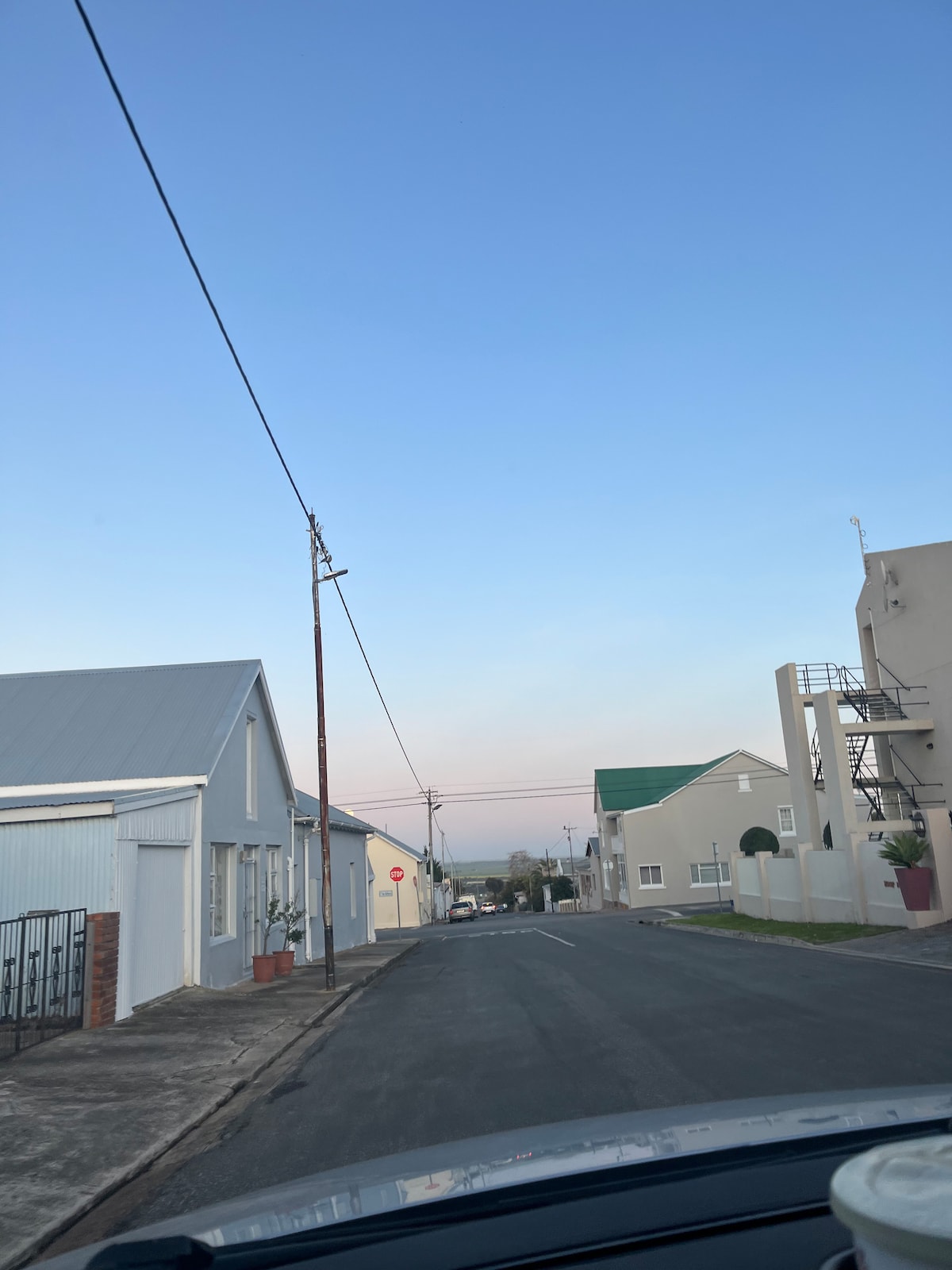 A quiet street is displayed, flanked by simple residential buildings with sloped roofs. Utility poles line the road, and a clear blue sky is visible above. Soft colors of dusk are hinted in the distance, suggesting a peaceful suburban environment.