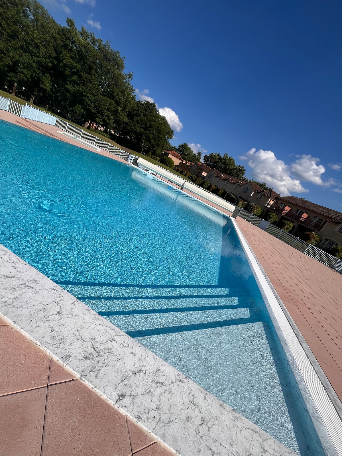 The image shows a spacious outdoor swimming pool with clear blue water. The pool's steps are visible in the foreground, leading into the deep end. Surrounding greenery and residential buildings can be seen in the background, complementing the pool area.