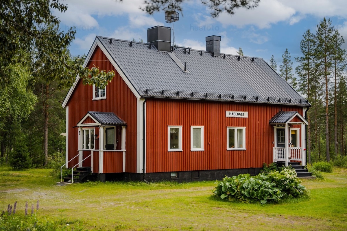 A charming red wooden station house is depicted, featuring a sloped roof and two entrances. Surrounded by greenery, the house is situated in a tranquil natural setting with tall trees in the background, emphasizing its inviting outdoor space.