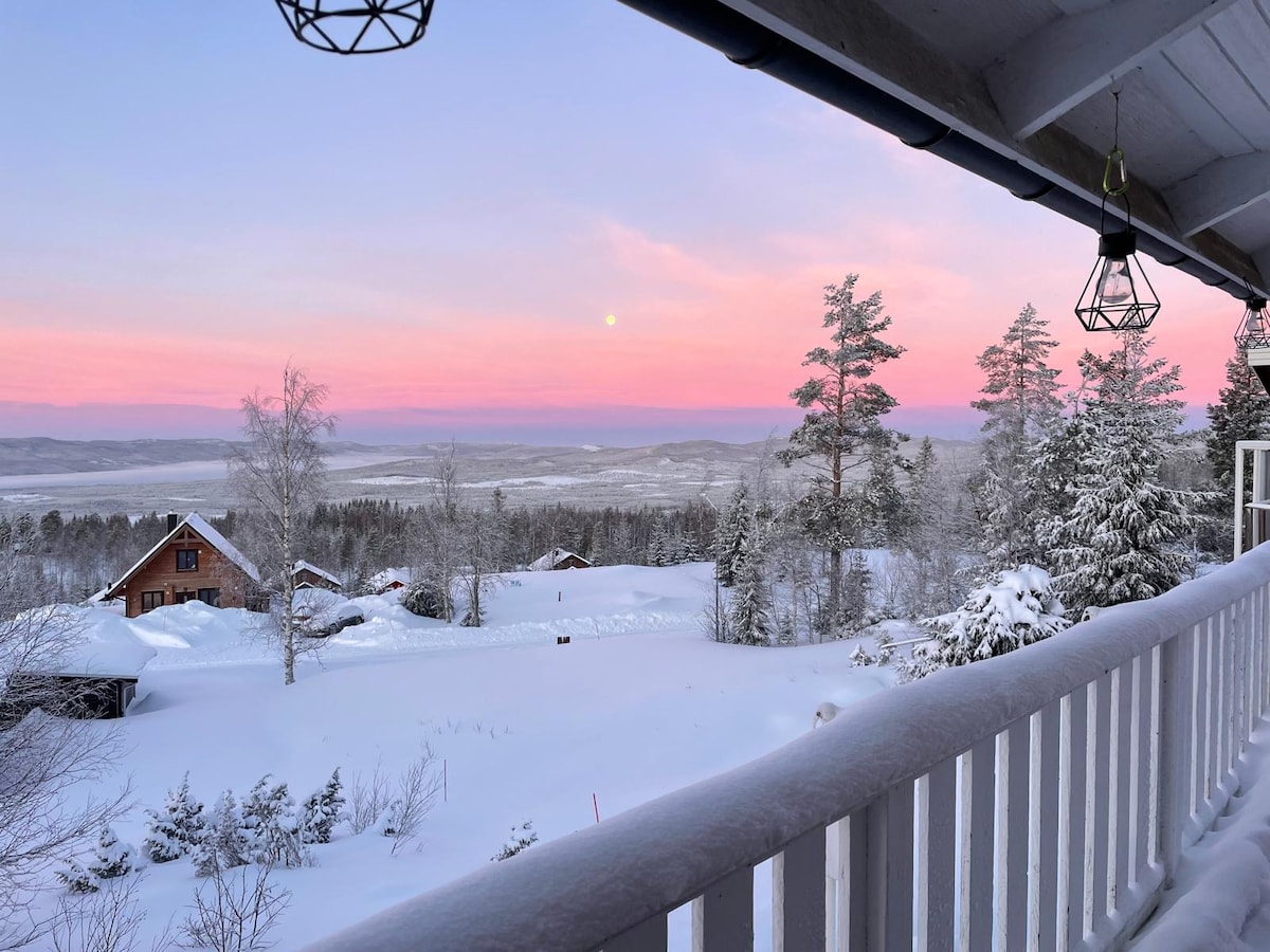 A serene winter landscape is viewed from a balcony, featuring a blanket of snow covering the ground and trees. The sky is illuminated in soft pink and purple hues, with a faint sun visible on the horizon. Nearby quaint cabins are partially hidden by snow.