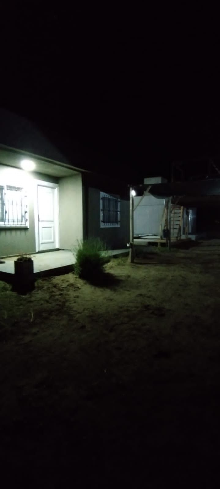 A well-lit entryway is visible, featuring a white door with a window. The surrounding area is landscaped with grass and small plants, while a shadowed structure is partially seen in the background. Soft lighting enhances visibility in the evening setting.