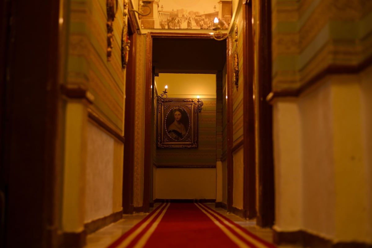 A narrow hallway is framed by decorative walls featuring intricate patterns. Dim lighting creates a warm ambiance, while plush carpeting with red and yellow stripes runs along the center. Framed artwork is displayed on the walls, adding character to the serene passageway.