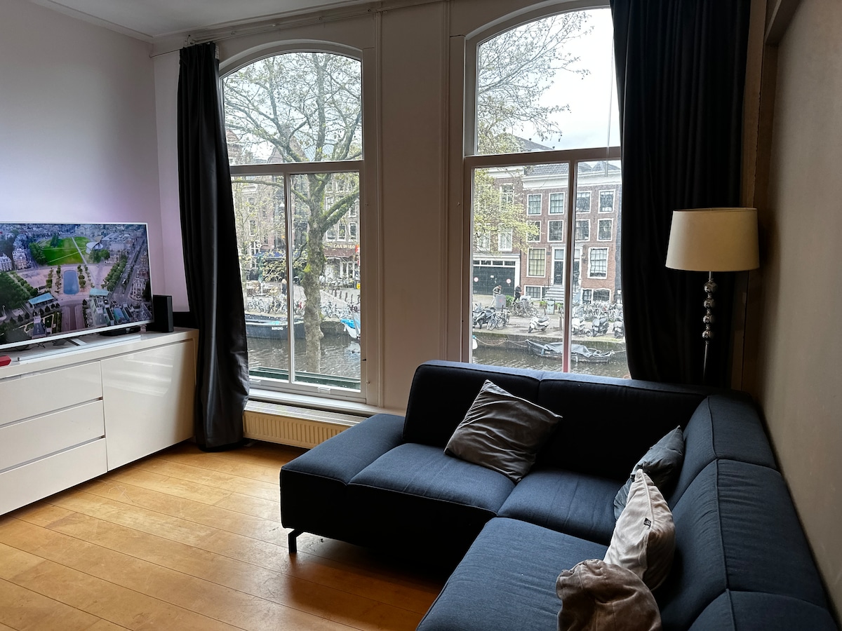 A comfortable living area features a deep blue sectional couch adorned with decorative cushions. Large windows allow natural light to fill the space, providing views of the picturesque canals and nearby architecture. A flat-screen TV is positioned on a sleek white media console.