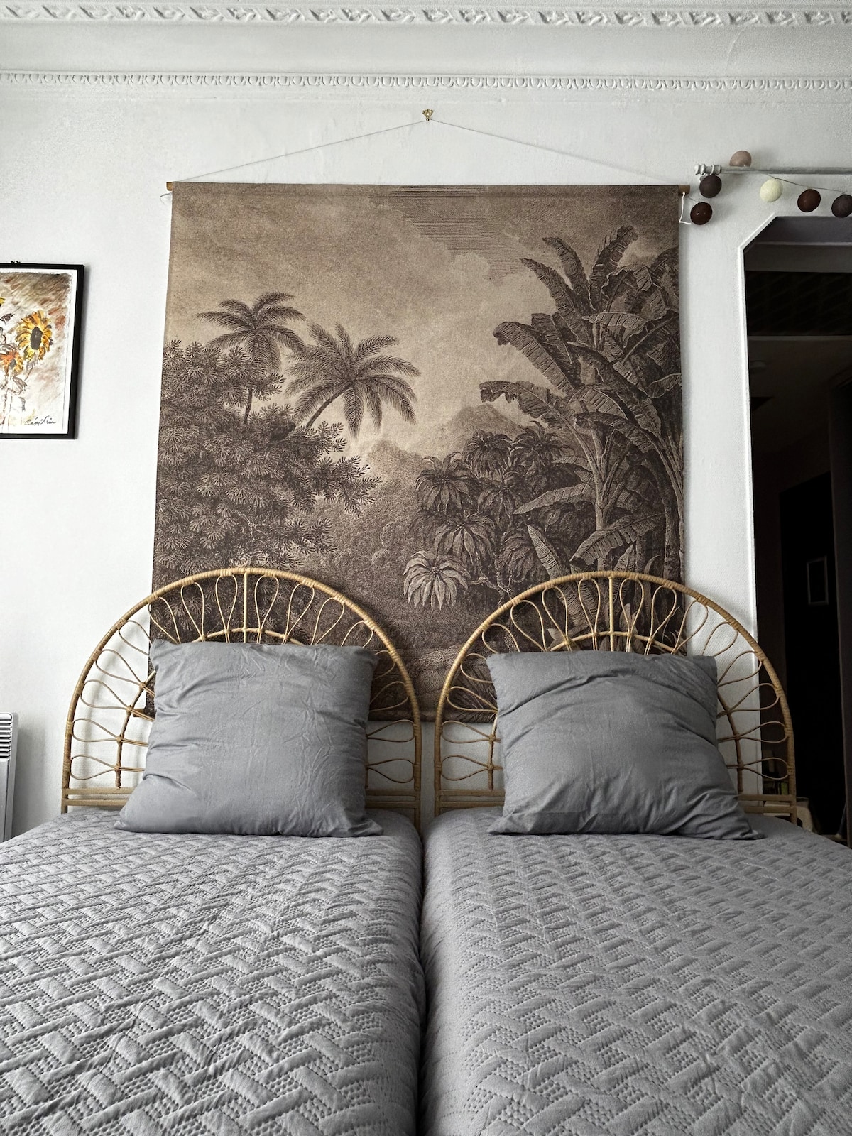 The sleeping area features two single beds arranged side by side, each adorned with gray pillowcases. A tapestry depicting a lush landscape hangs above the beds. The walls are decorated with framed artwork, contributing to the room's overall aesthetic.