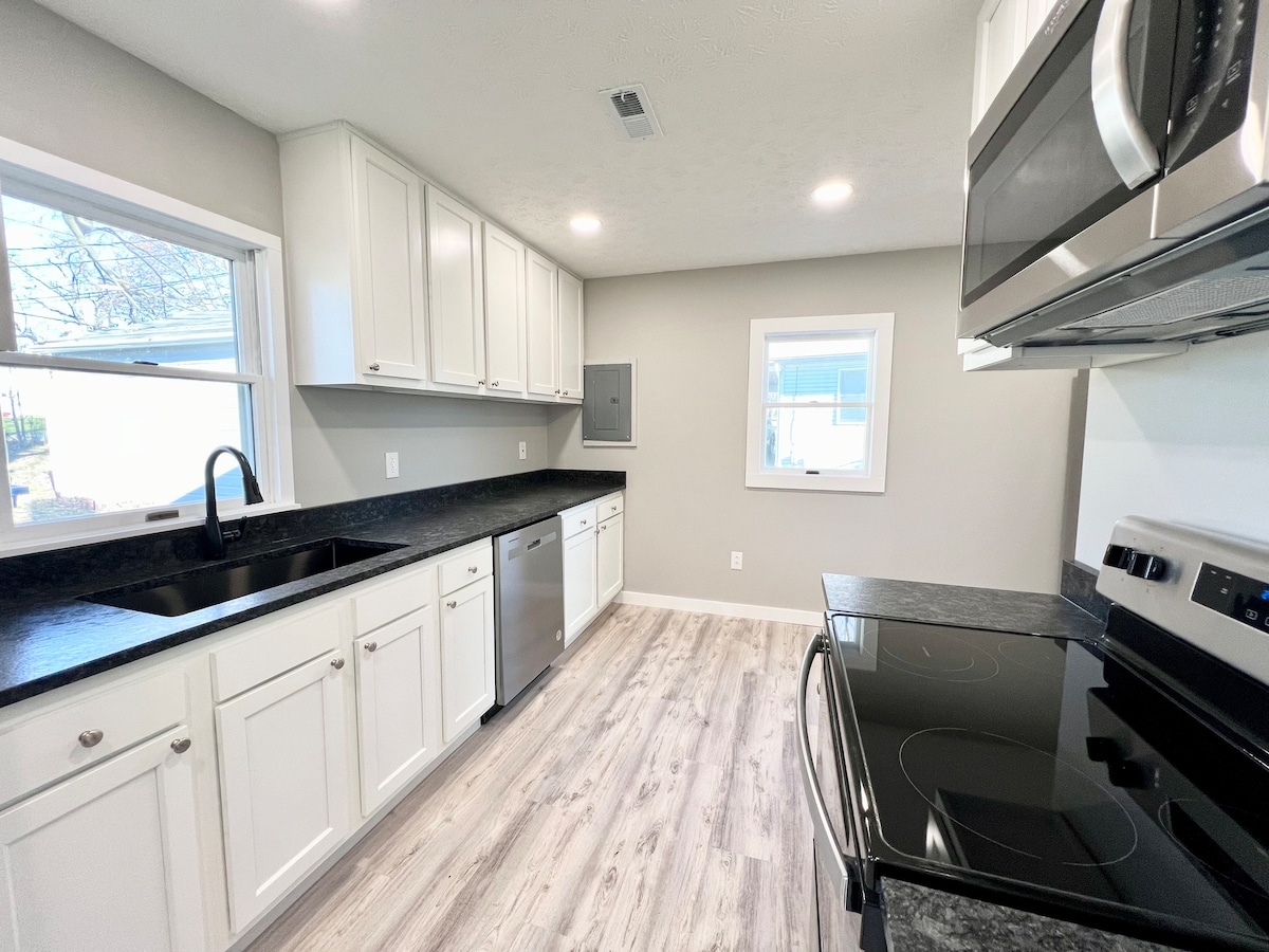 The kitchen showcases modern cabinetry with a dark countertop. Stainless steel appliances, including a microwave and oven, are positioned alongside a window that allows natural light to brighten the space. The flooring features a light wood finish, contributing to a clean and updated look.