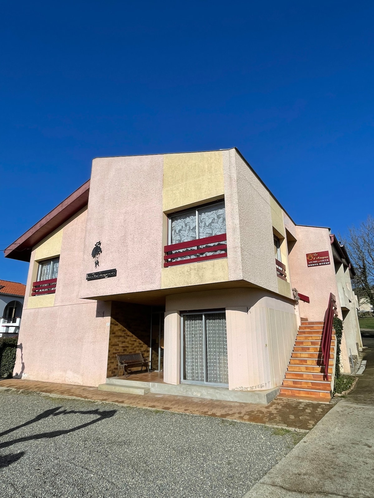 The exterior of the building displays a two-story structure with a blend of pastel pink and beige colors. Decorative detailing is visible around the windows, while steps lead to the entrance. A clear blue sky serves as the backdrop, enhancing the overall appearance.