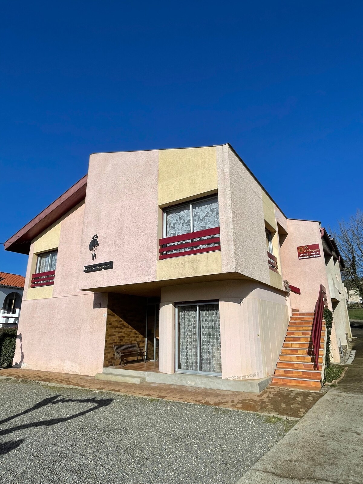The building features a two-story structure with a light pink facade and stone accents. Balconies with maroon railings are visible on both levels. A staircase leads to the entrance, framed by a grassy area and clear blue sky.