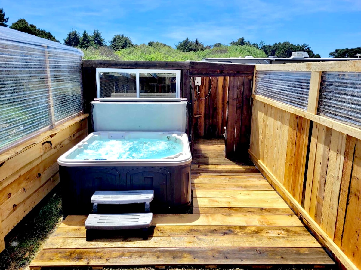 An outdoor hot tub is situated on a wooden deck, enclosed by wooden fencing and clear panels. Sunlight illuminates the area, enhancing the tranquil setting. A shower is visible nearby, and the surrounding greenery adds to the natural ambiance.