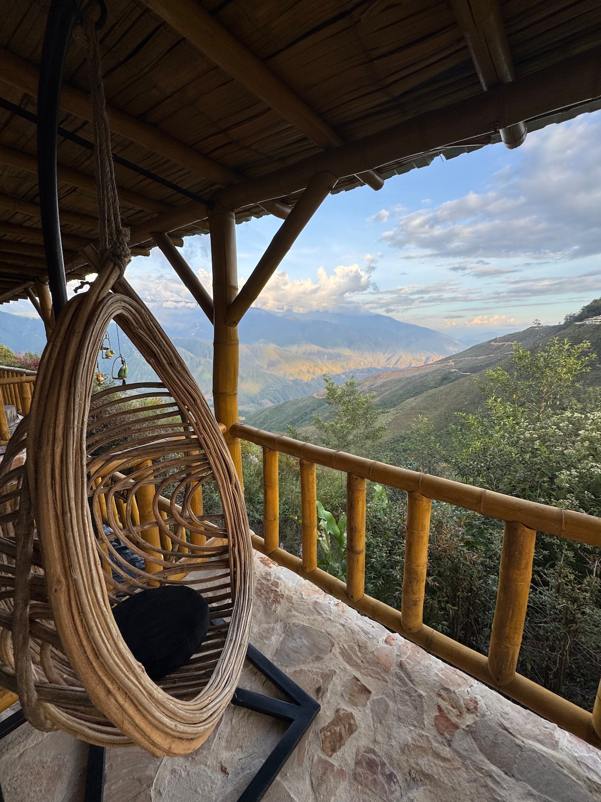 A uniquely designed hanging chair crafted from natural materials is positioned on a covered patio. Views of rolling hills and distant mountains are visible in the background, framed by lush greenery and a clear sky.