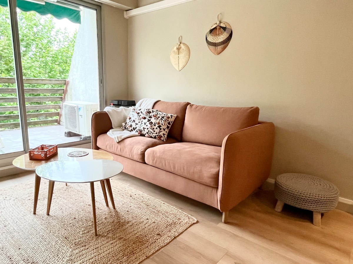 A cozy living area features a soft, light-colored sofa adorned with patterned cushions. A round coffee table occupies the center, surrounded by a textured area rug. Natural light comes through a large window, with greenery visible outside, enhancing the serene atmosphere.