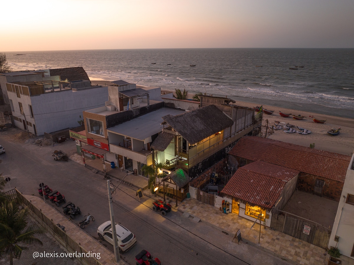 An aerial view captures a coastal area at sunset, showcasing a beachside establishment with a thatched roof. The shoreline is visible, lined with fishing boats, while the surrounding streets display parked vehicles. The warm hues of the sunset reflect on the water, enhancing the seaside ambiance.