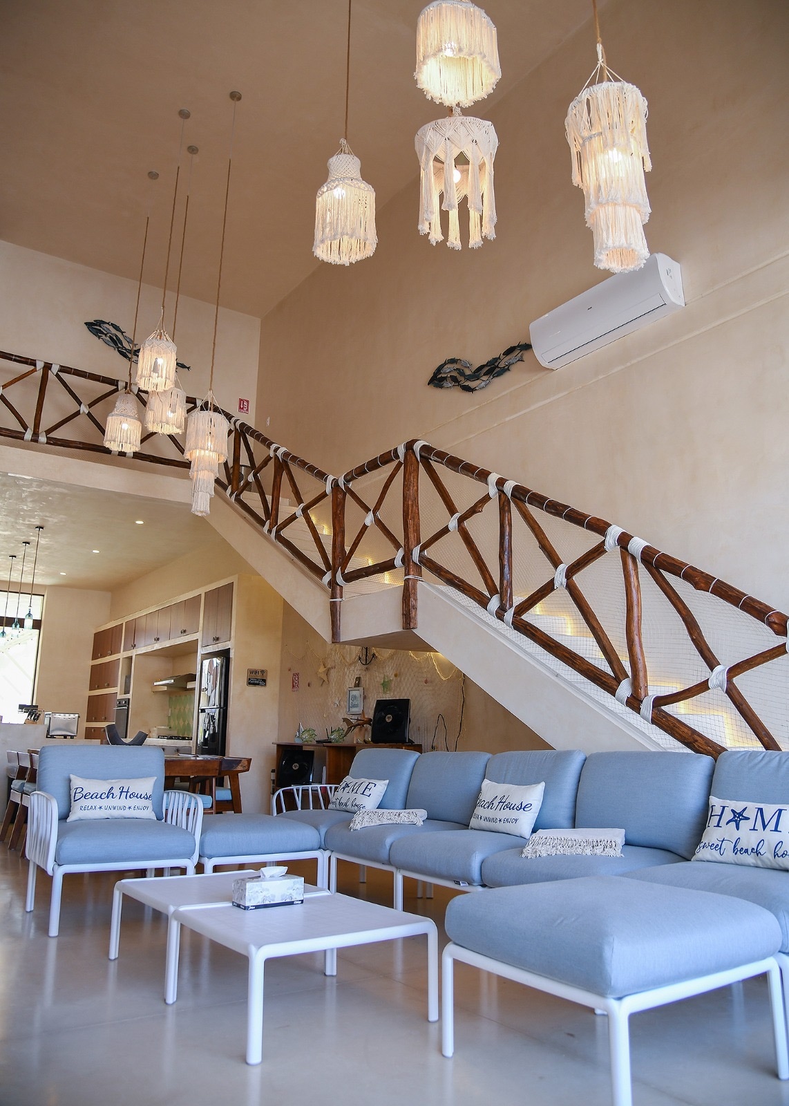 An open living area features a relaxed seating arrangement with light blue couches adorned with decorative pillows. A staircase with wooden railing is visible in the background, complemented by hanging light fixtures. The space is designed with neutral tones and contemporary decor.