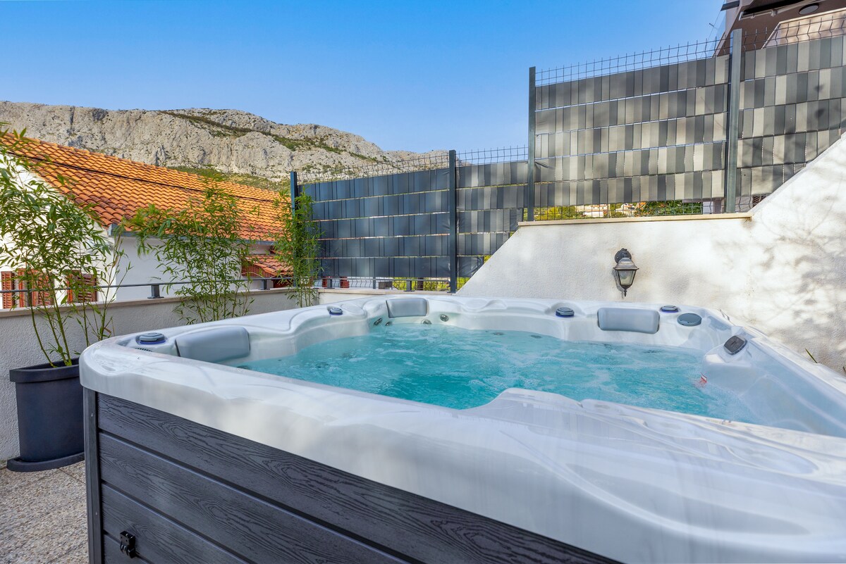 A heated jacuzzi is prominently featured, surrounded by a secure enclosure. The clear blue water contrasts with the natural landscape of mountains in the background. Planting and fencing enhance the sense of privacy in this outdoor relaxation area.