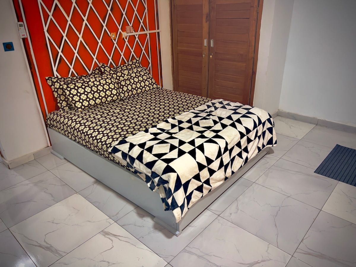 A spacious bed is dressed in a patterned black and white blanket, complemented by decorative pillows. The accent wall behind the bed features a bright orange hue. Flooring consists of glossy white tiles, with a soft mat placed beside the bed.
