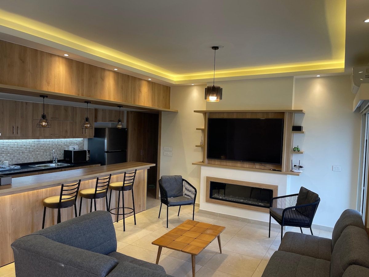 A modern living area showcases comfortable seating arrangements, including a gray sofa and stylish chairs. A wooden coffee table sits at the center. The kitchen bar with high stools is visible behind, and a large flat-screen television is mounted on the wall alongside a contemporary fireplace.