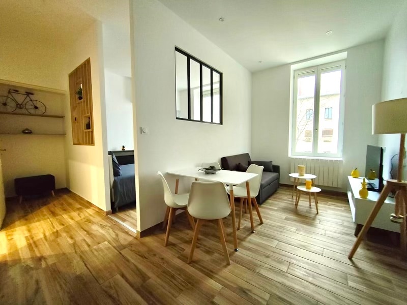 A modern living space is presented with light wood flooring and bright walls. A small dining table with white chairs is visible, alongside a gray sofa. Natural light filters through a large window, enhancing the airy feel. A partition leading to a bedroom area is partially shown.