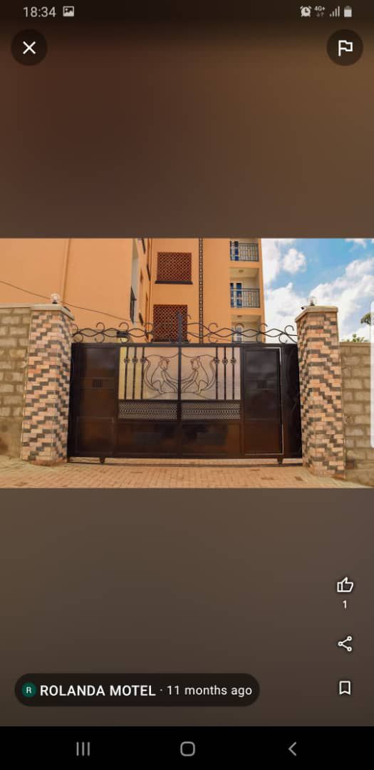 A wrought iron gate displays an intricate design of two figures in a welcoming pose, flanked by textured stone pillars. The surrounding walls are light gray with decorative brick patterns, creating a distinctive entrance to the property.