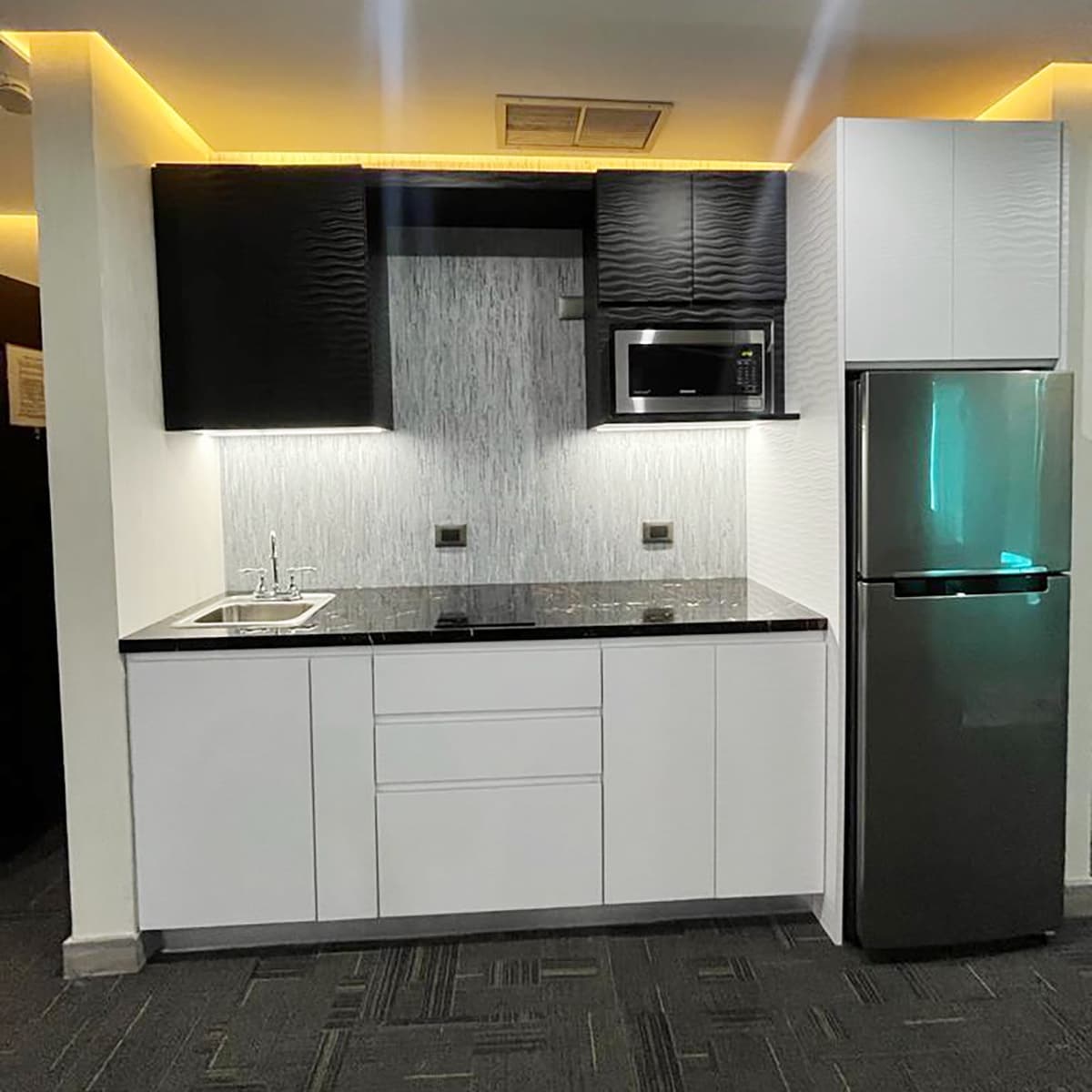 A modern kitchenette features sleek black and white cabinetry, with a stainless steel refrigerator and microwave. A small sink is set into a dark countertop, while under-cabinet lighting provides a soft glow over the space. Gray patterned flooring complements the design.
