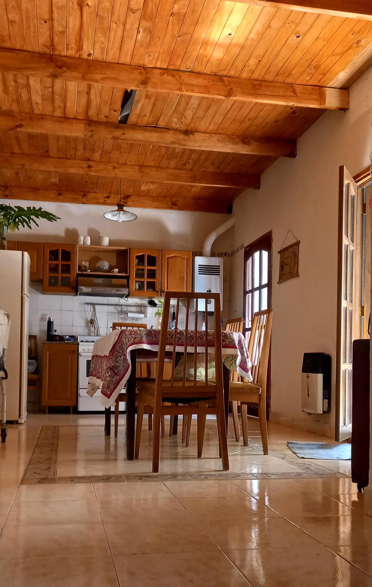 A welcoming dining area is highlighted by a wooden table surrounded by six chairs. Natural light fills the room through glass doors, reflecting off the tiled floors. The ceiling showcases exposed wooden beams, and kitchen amenities are visible in the background.