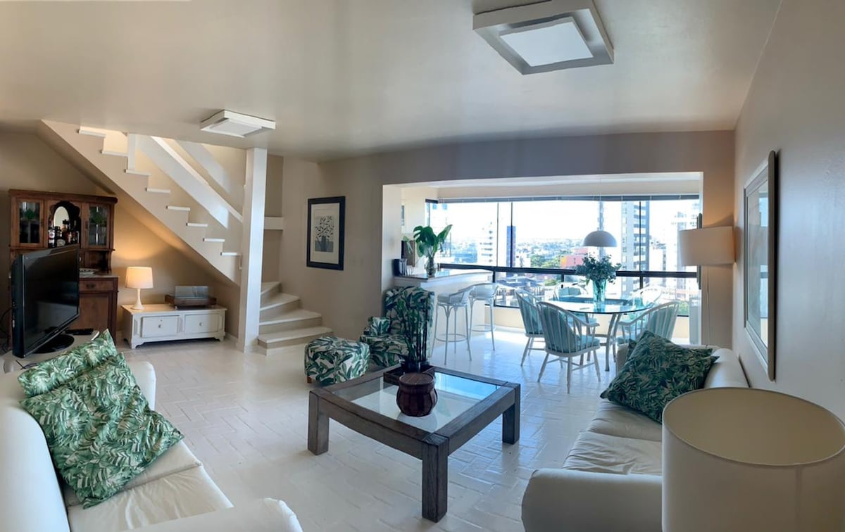 A spacious living area is filled with natural light, featuring comfortable seating arranged around a rectangular table. A glass window offers city views, and dining chairs are set around a round table in the background. Decorative plants add a touch of greenery.