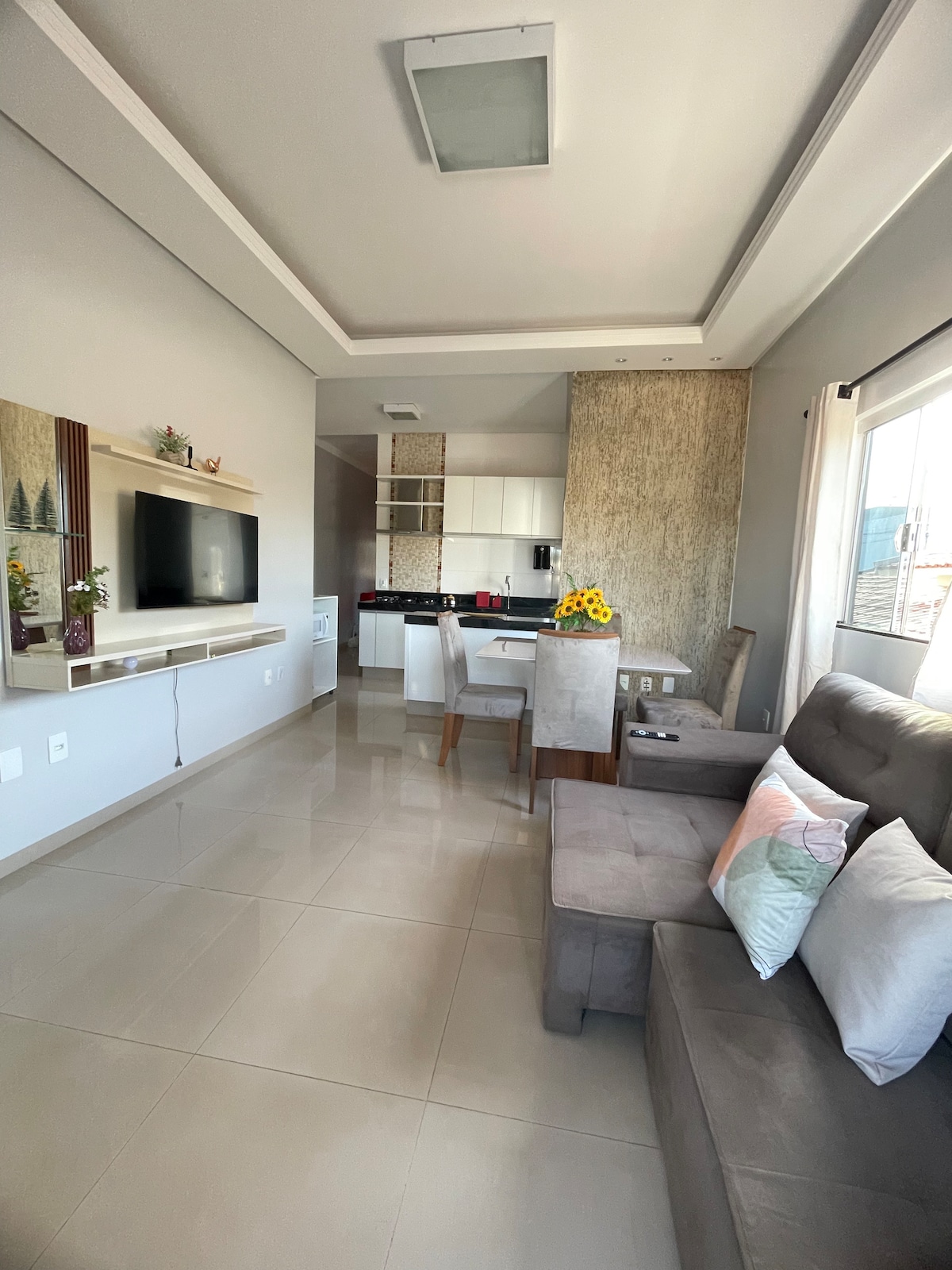 A modern living area features a cozy gray sofa adorned with decorative pillows. A small dining table is set nearby, with natural light streaming through a large window. The open kitchen design integrates seamlessly with the space, showcasing sleek cabinetry and a subtle color palette.