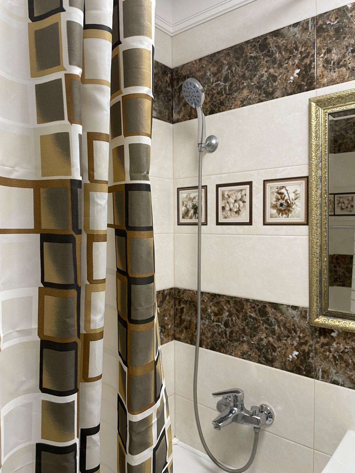 A compact bathroom features a shower with a modern handheld faucet and decorative tile accents. A patterned shower curtain adds texture, while a framed mirror reflects the space. The area showcases a blend of neutral and gold tones on the walls and fixtures.