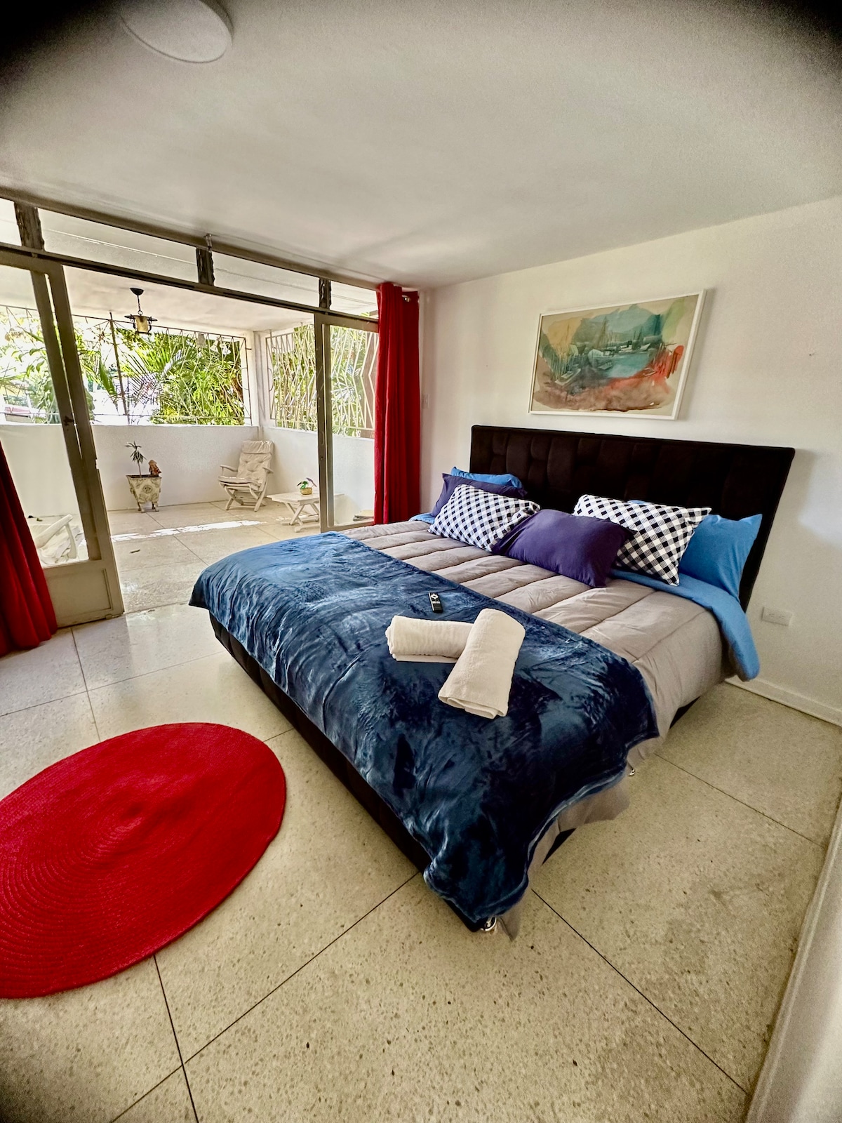 A spacious bedroom features a large bed with a navy blue blanket and patterned pillows. Natural light streams through glass doors leading to a private terrace with seating. A vibrant red rug lays on the floor, complementing the modern decor and serene atmosphere.