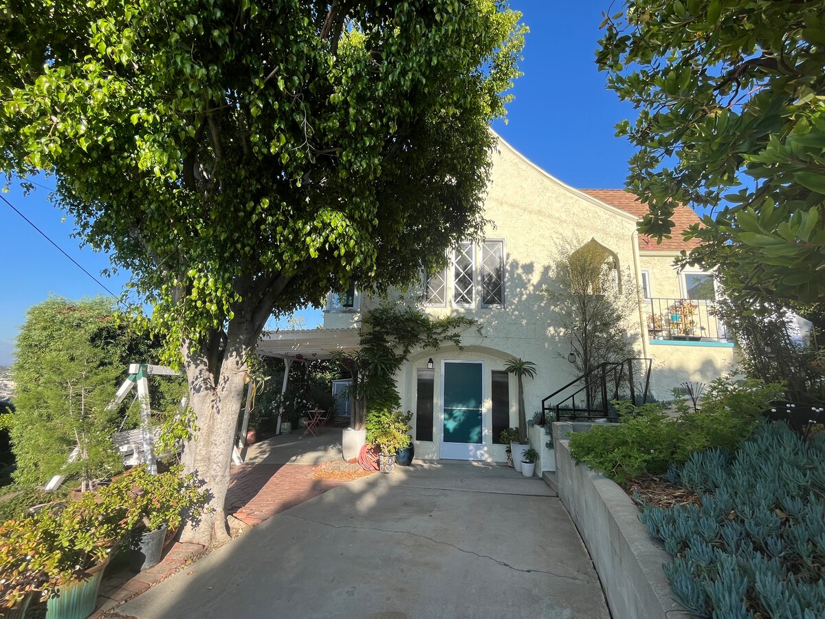 A charming exterior view of a hillside home is shown, featuring a wide driveway flanked by lush greenery. A large tree provides shade, while the entrance boasts a set of steps leading to a blue door, complemented by decorative plants and a sunny sky.