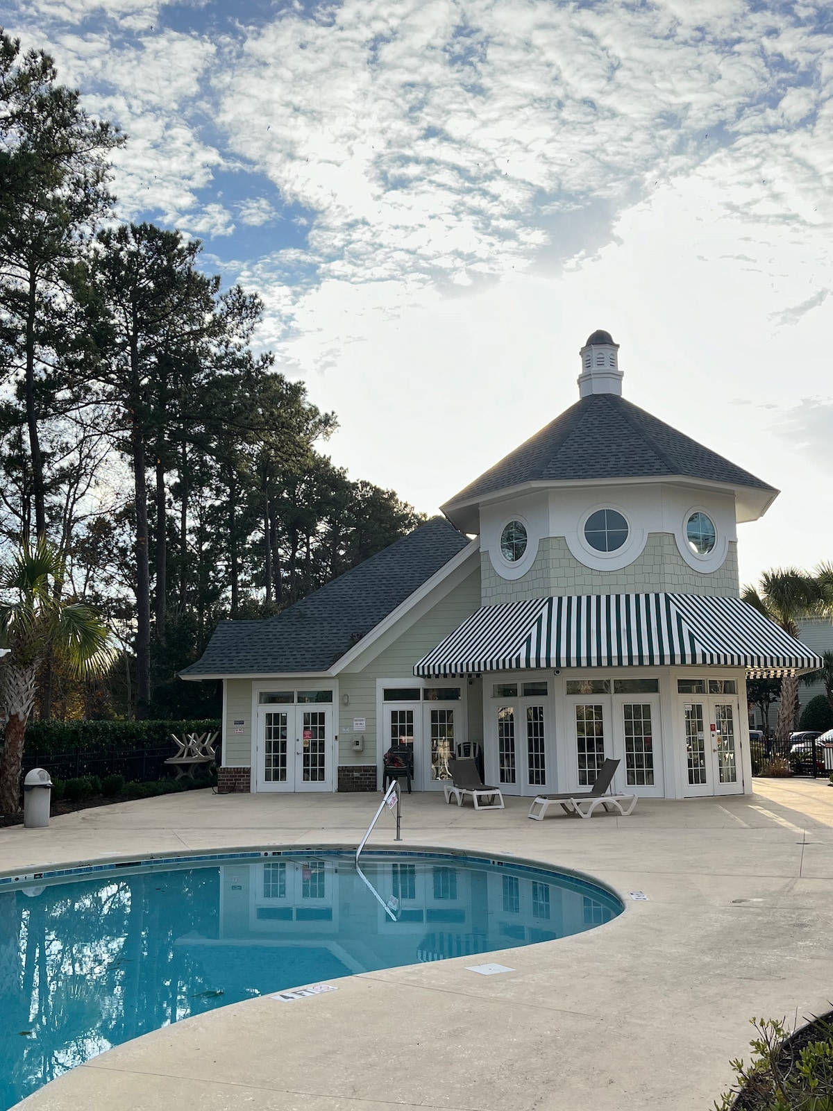 A charming clubhouse features large windows showcasing a circular design, topped with a conical roof. The inviting pool area is surrounded by lounge chairs, while a striped awning provides shade. Lush greenery adds to the serene ambiance of the space.