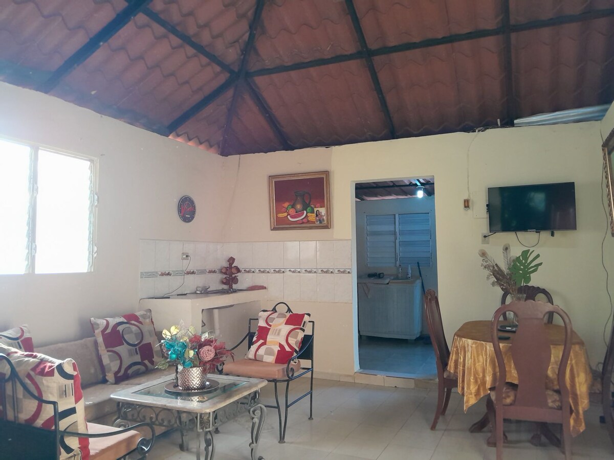 The interior space features a cozy seating area with patterned cushions arranged around a low table. A dining area with a table and chairs is visible, and a small kitchen area can be seen in the background. Natural light enters through a window, highlighting the warm color palette.