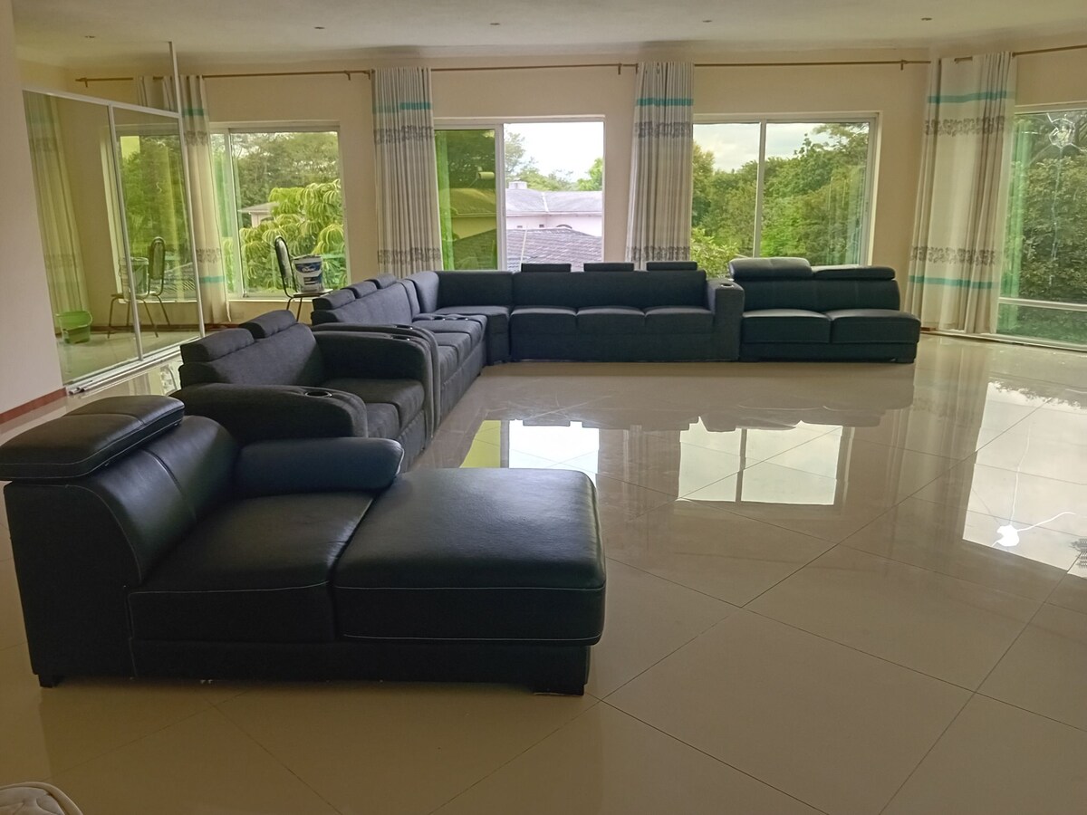 A spacious living area features a large sectional sofa arranged to create a social atmosphere. Floor-to-ceiling windows allow natural light to flood in, offering views of the surrounding greenery. The polished tile floor reflects the ample light, enhancing a sense of openness.