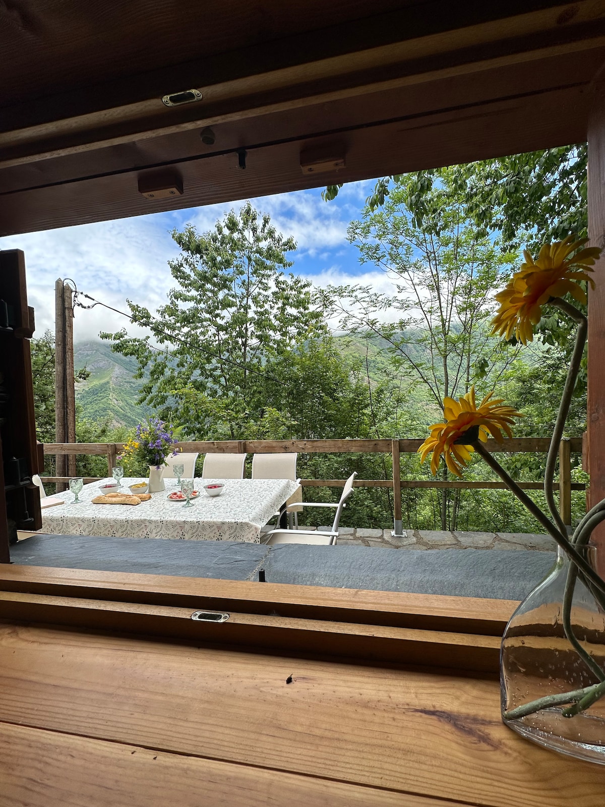 A view from a wooden window frame highlights an outdoor dining area set on a deck. A table is adorned with a tablecloth and surrounded by chairs, while greenery and a distant mountain vista create a serene backdrop. Sunlight filters through the trees.