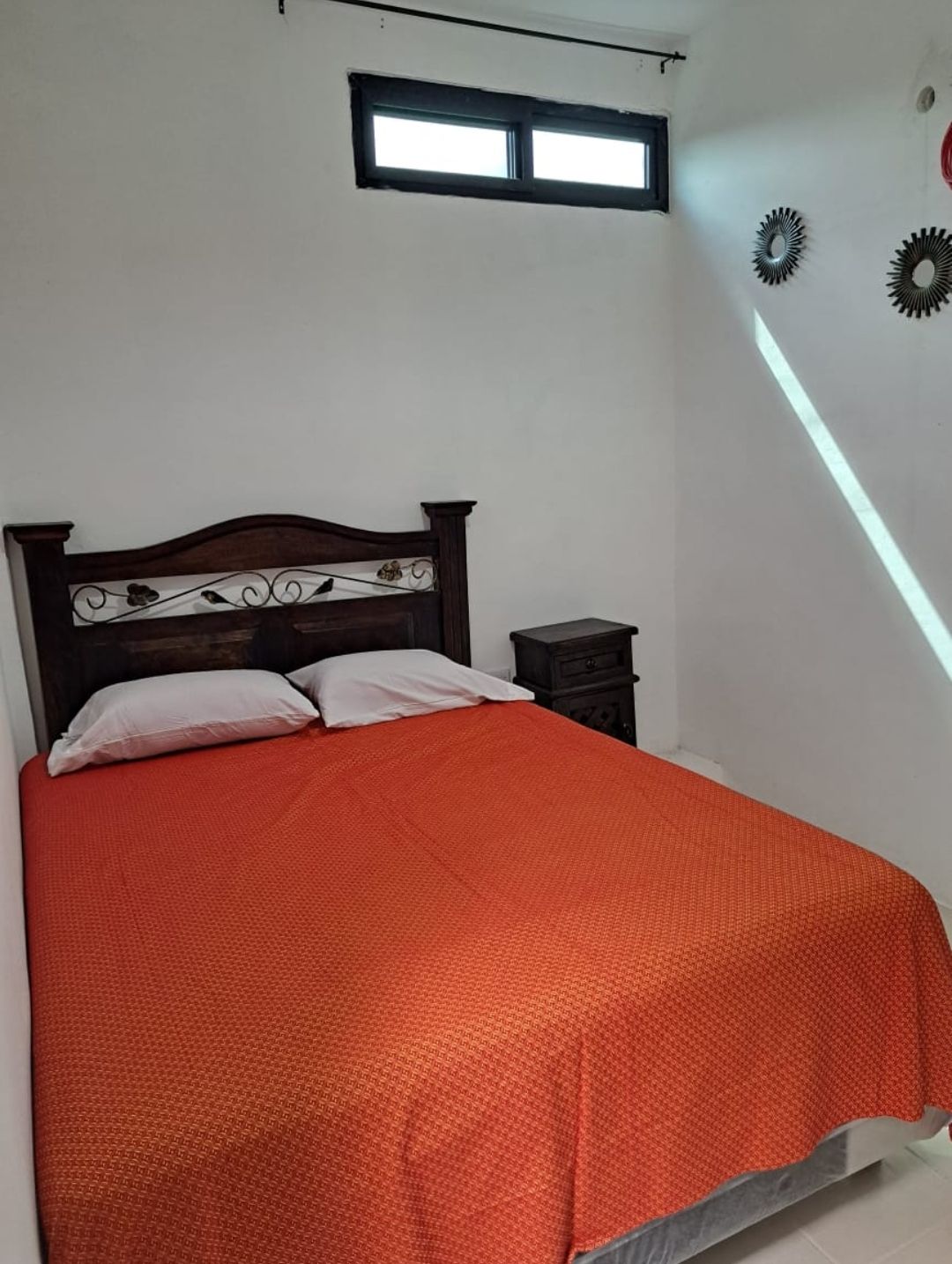 A sleeping area features a wooden headboard with decorative elements. An orange bedspread covers the spacious bed, complemented by two white pillows. Natural light enters through a small window, creating a soft, warm glow in the room.