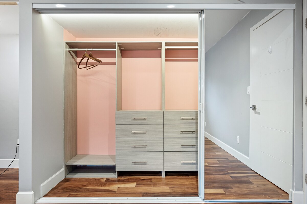 A spacious closet area is presented with a modern design. It features built-in drawers and a hanging space, complemented by soft lighting. The walls are painted in a light pink hue, contrasting with the natural wood floor visible in the adjacent area.