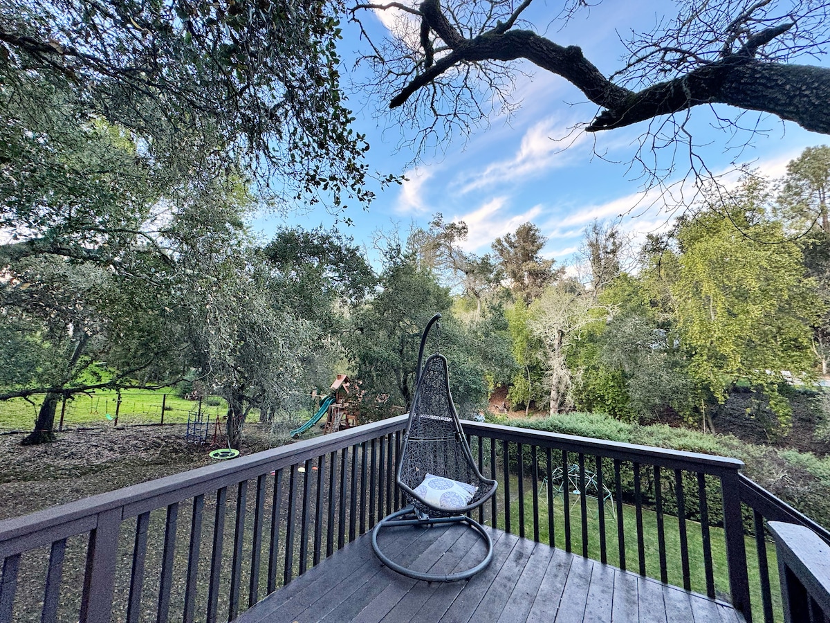 A private patio area with wooden decking is presented, featuring a unique hanging chair. Surrounding greenery includes trees and shrubs, enhancing the natural setting. The sky above shows scattered clouds, creating a serene outdoor space for relaxation.