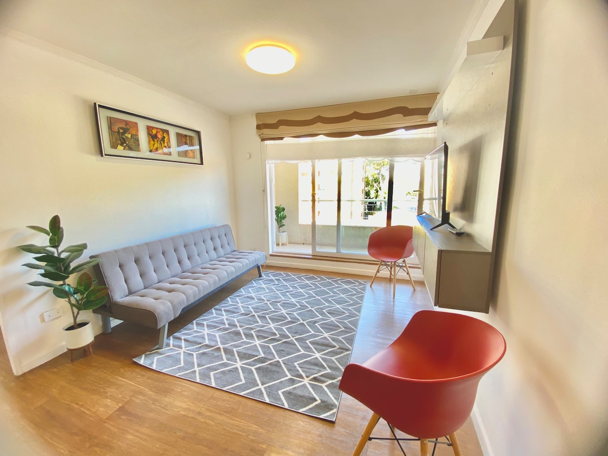 A bright living area features a gray sofa that accommodates multiple guests, positioned across from a wall-mounted television. Two red chairs offer additional seating, while a decorative rug adds texture to the wood floor. Large glass doors invite natural light and provide access to an outdoor space.