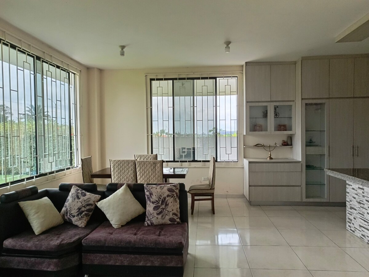 A modern living area featuring a dark sectional sofa adorned with decorative pillows. A bright dining table is positioned nearby, surrounded by light-colored chairs. Large windows allow natural light to fill the space, emphasizing the neutral tones of the interior.