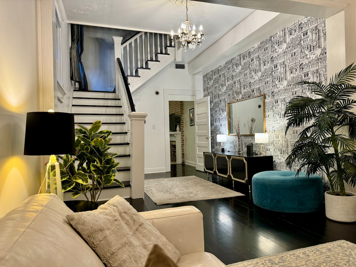 A well-appointed foyer showcases a light-colored sofa facing a plush ottoman. A decorative wall displays intricate patterns, while a stylish chandelier illuminates the space. A staircase with contrasting black and white steps leads to the upper floor, complemented by potted plants for a touch of greenery.