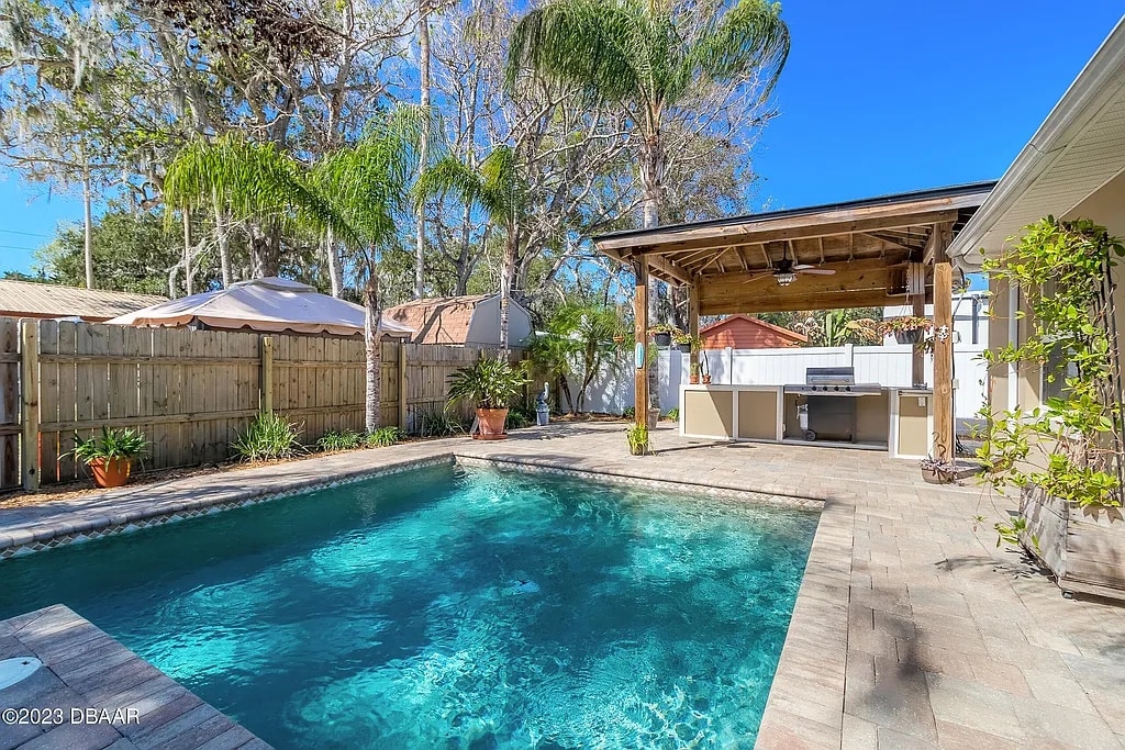A refreshing swimming pool is surrounded by paved decking and lush greenery. A shaded outdoor kitchen area features a countertop and overhead fan. Various plants and landscaping elements create a natural ambiance, while the clear blue sky enhances the outdoor setting.