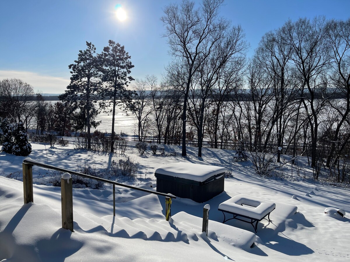A serene winter landscape is displayed, featuring a snow-covered yard that leads to a glimmering lake. Bare trees stand against a clear blue sky, while sunlight reflects off the water's surface. A hot tub is visible near the snow-draped steps.