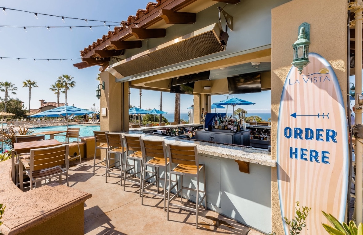 An open-air bar area is highlighted under a clear blue sky, featuring high-backed wooden stools arranged around a granite countertop. On the right, a surfboard sign points towards the bar with 'ORDER HERE' written on it. Umbrellas provide shade over the seating and pool area in the background.